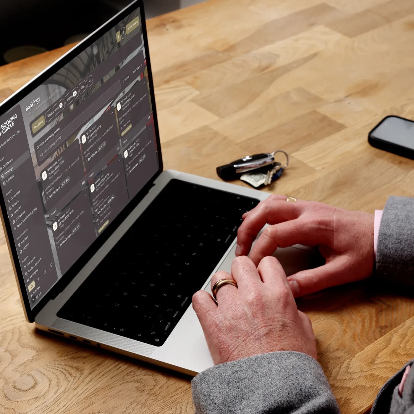 Hands on a laptop showing the Booking Circle bookings list, on a wooden desk with car keys and a phone beside it.