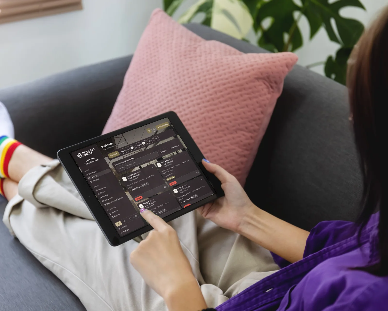 A woman lying back on a sofa, tapping a tablet that shows the Booking Circle bookings list.